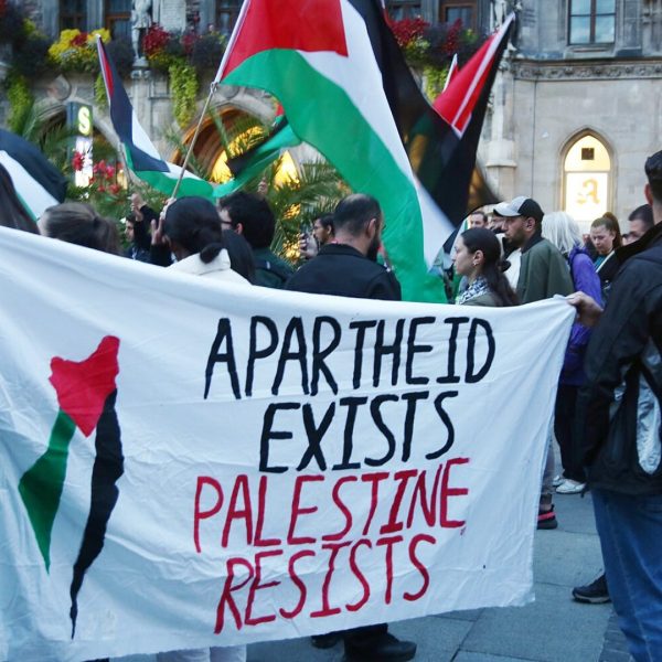 People in the streets wave Palestinian flags and hold a banner that reads, APARTHEID EXISTS PALESTINE RESISTS.